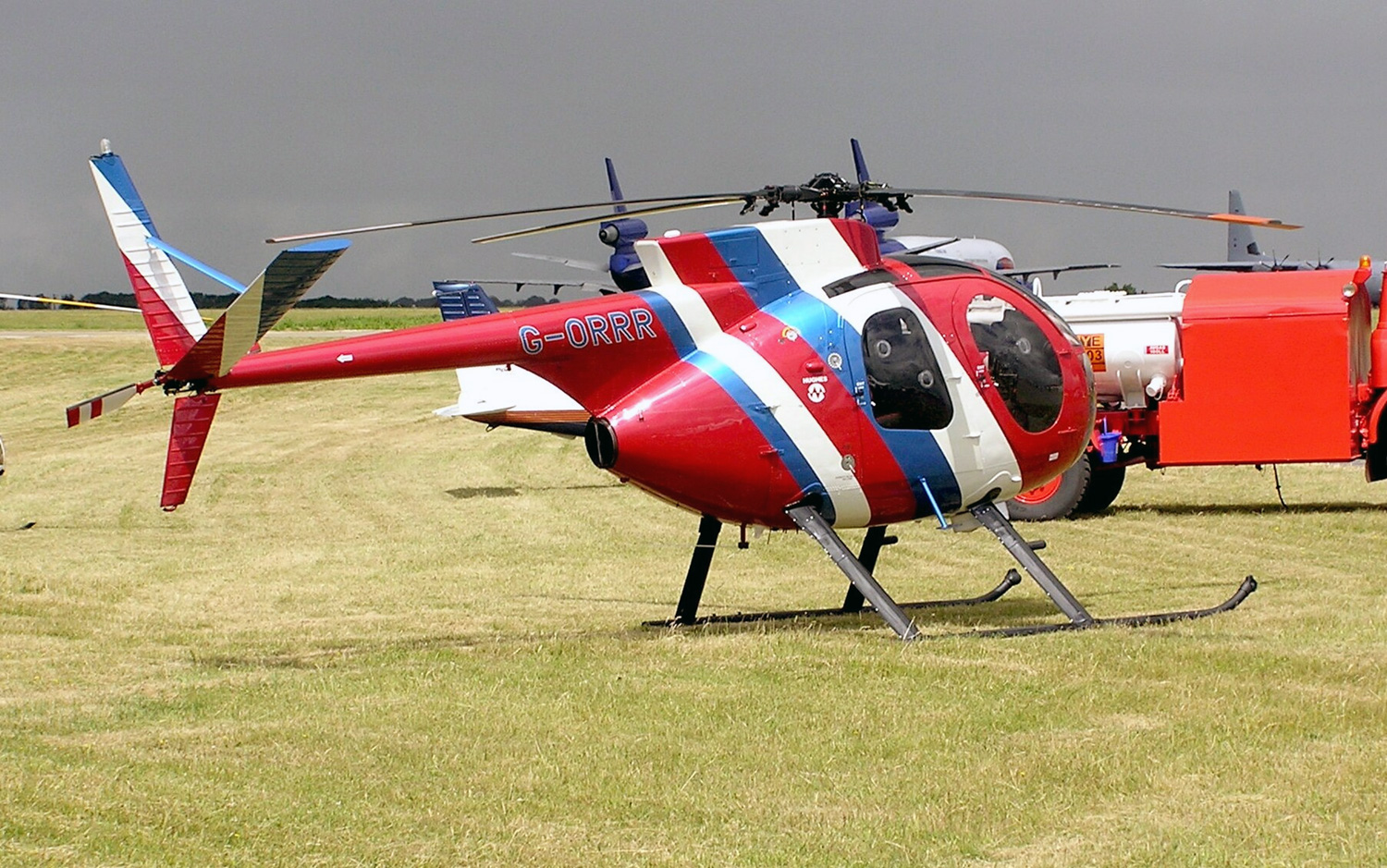 Photo of a red, white, and blue Hughes 500 setting in a grass field under bright sunshine