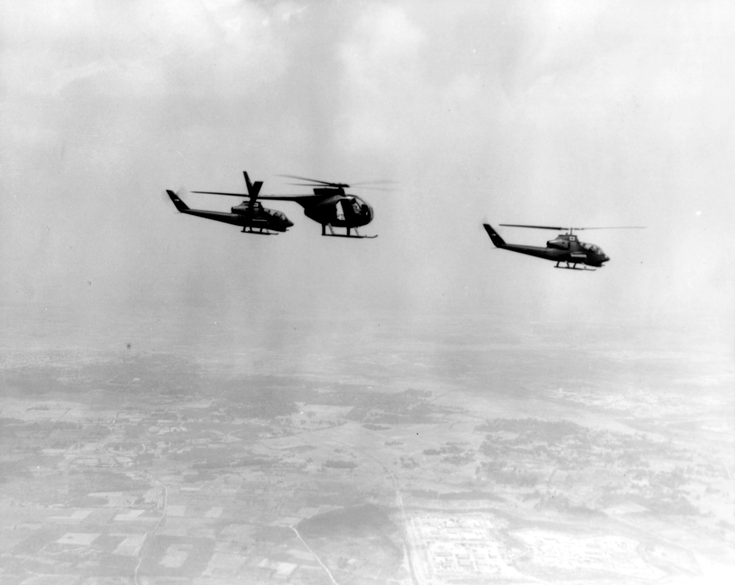 High altitude B&W photo of a Hughes 500 as OH-6A in formation with a pair of AH-1G Cobra attack helis flying left to right at altitude during the Vietnam War era.